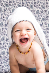 malabar baby white bamboo cotton towel shown on a cute and happy baby. The towel is white and silky soft and has a hood. The towel also has a detailed pom pom trim. The towel is extra large and fits newborns - toddlers 5 years old. Pom Pom colors are shown in neutral grey. The happy baby is also laying on malabar baby's Provence blue cotton crib quilt.