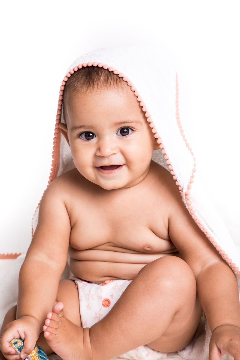 malabar baby white bamboo cotton towel shown on a cute and happy baby. The towel is white and silky soft and has a hood. The towel also has a detailed pom pom trim. The towel is extra large and fits newborns - toddlers 5 years old. Pom Pom colors are shown in sweet peach.